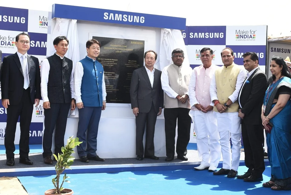 Officials in suits and kurtas line up beside an unveiled plaque in front of Samsung and Make in India banners, with a potted sapling in the foreground.