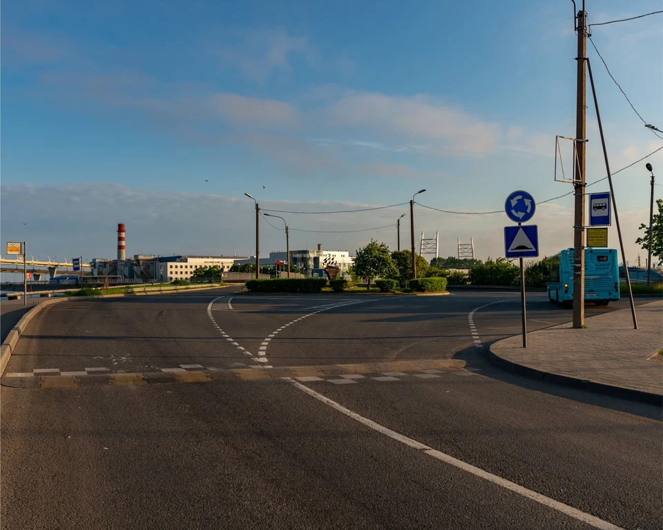 Empty road junction on a clear evening, with lane markings, roundabout and pedestrian-crossing signs, a bus shelter, street lights and low buildings in the distance.