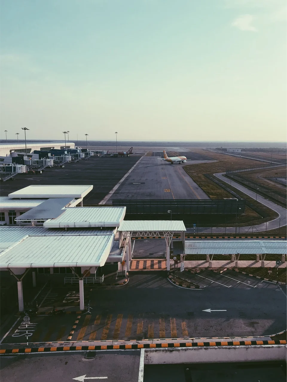 Elevated view across an airport, with the white canopy roofs of a terminal forecourt in front and a long runway stretching away to a lone aircraft under a hazy sky.