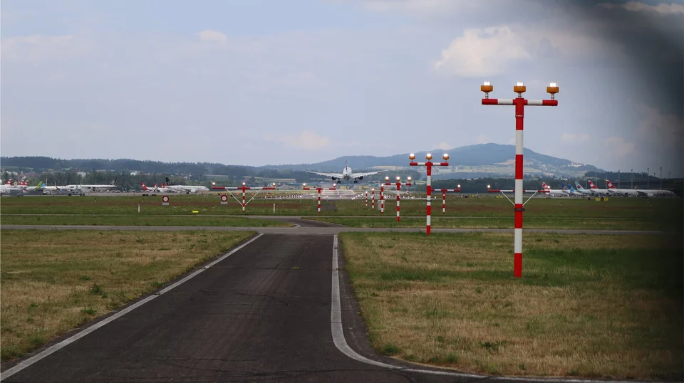 View along a service road towards an airport runway, with red-and-white approach light masts on the grass and a plane on the runway against distant hills.