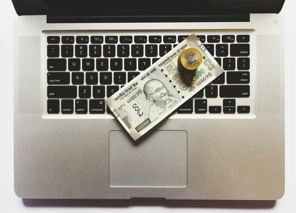 An Indian 500-rupee note and a small stack of coins resting on the keyboard of a silver laptop, seen from above.
