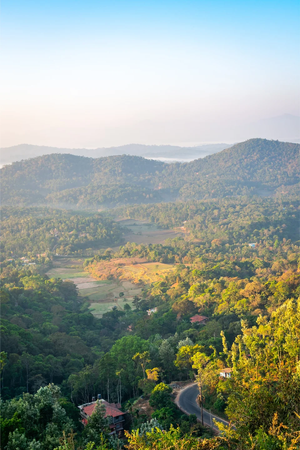 Looking down over forested hills and a green valley under a hazy sky, with a few red-roofed houses and a road winding through the trees.