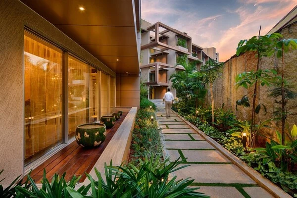 A stone-paved garden path lined with tropical plants runs beside a glass-fronted building with a wooden deck and planters, as a man walks towards a terraced block at dusk.