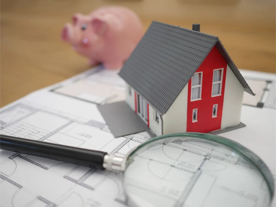 Small model house with a red front and grey roof standing on architectural floor plans, with a magnifying glass in front and a pink piggy bank behind.
