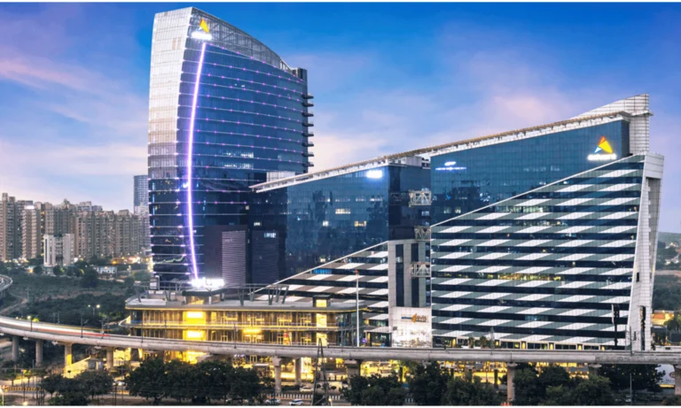 Glass-fronted office towers lit up at dusk, one with a curved facade and a vertical light strip, beside an elevated roadway with apartment blocks behind.