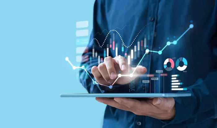 Person tapping a tablet as line graphs, candlestick charts and pie charts float above the screen.