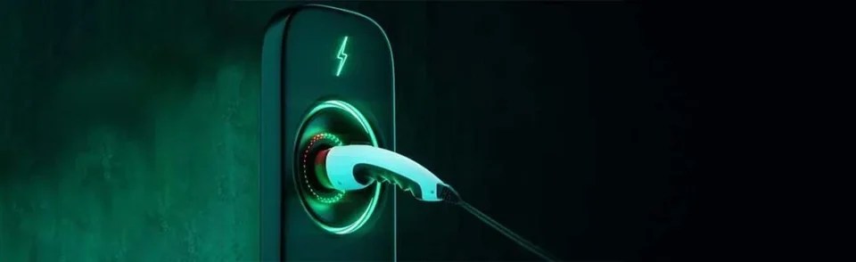 An electric vehicle charging cable plugged into a wall-mounted charger marked with a lightning bolt, lit in green against a dark background.